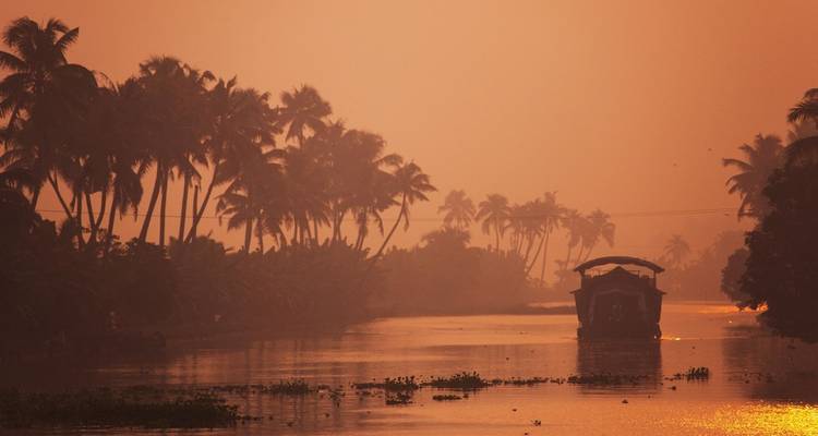 Silhouettes eines Hausbootes gleitet durch einen von Palmen gesäumten Kanal im warmen orange Licht der Morgenröte