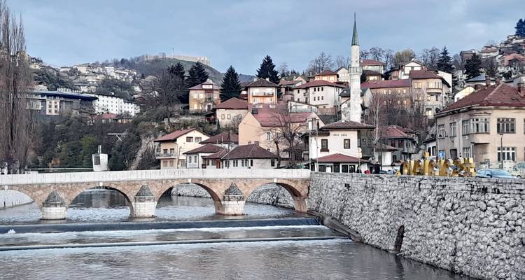 Un pont de pierre historique enjambant la rivière Miljacka avec des maisons construites sur les collines et un minaret élancé à Sarajevo.