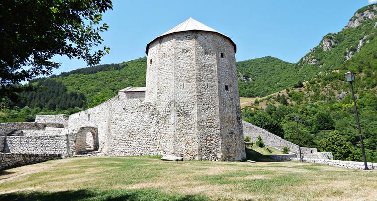 Une tour de pierre cylindrique et des murs de forteresse situés sur des terrains herbeux entourés de collines boisées.