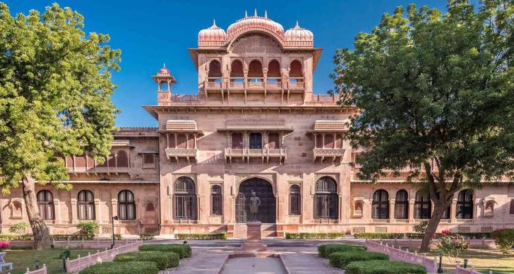 Elegant pink-sandstone Laxmi Niwas Palace framed by lush trees under a bright blue sky.