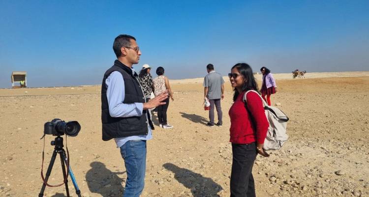 Un guide s'adresse aux visiteurs sur un plateau sablonneux près des pyramides, avec un appareil photo et un chameau visibles en arrière-plan.