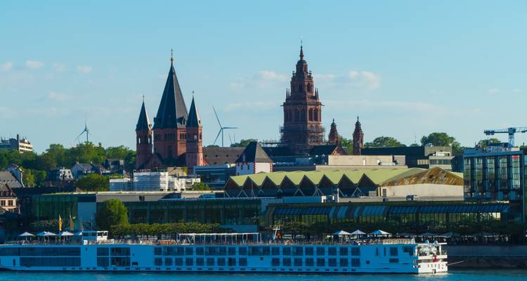 Panoramic view across the Rhine to Mainz with its red-brick cathedral spires, modern glass buildings and a white river cruise ship in the foreground.