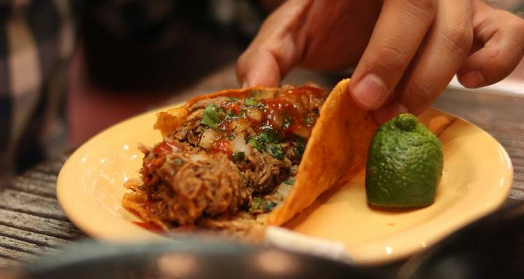 Hand hebt einen Taco mit zerkleinertem Fleisch, Salsa und Koriander neben einem Limettenschnitz auf einem Keramikteller.