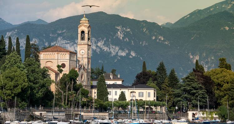 Peaceful lakeside Italian village with church tower against forested mountains and marina of sailboats.