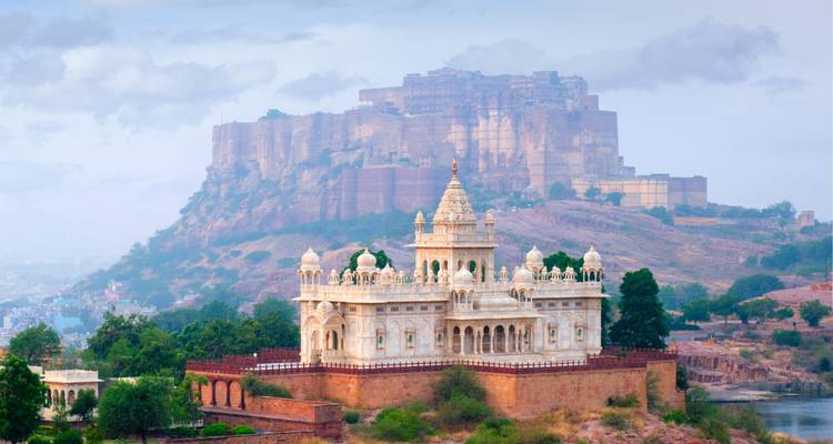 Elegant white marble memorial sits in front of a massive sandstone fort atop a hazy hill.