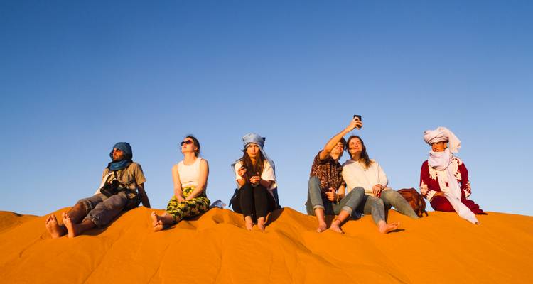 Grupo de turistas con pañuelos coloridos sentados en la cresta de una duna tomándose selfies al atardecer