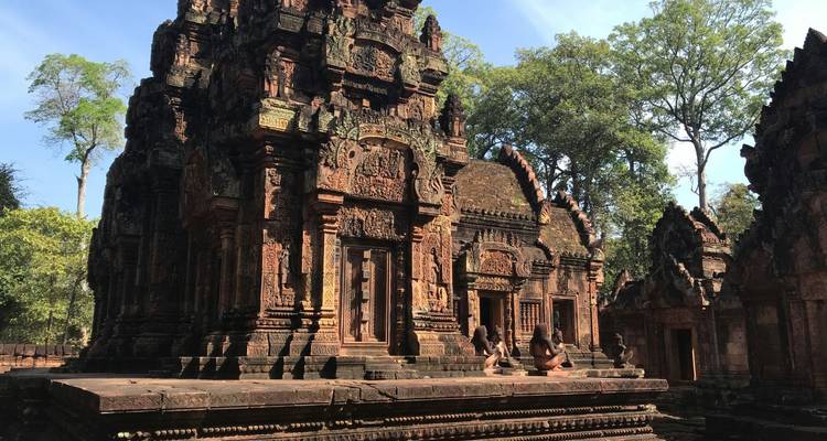 Intricately carved red sandstone temple of Banteay Srei with two visitors sitting on steps.