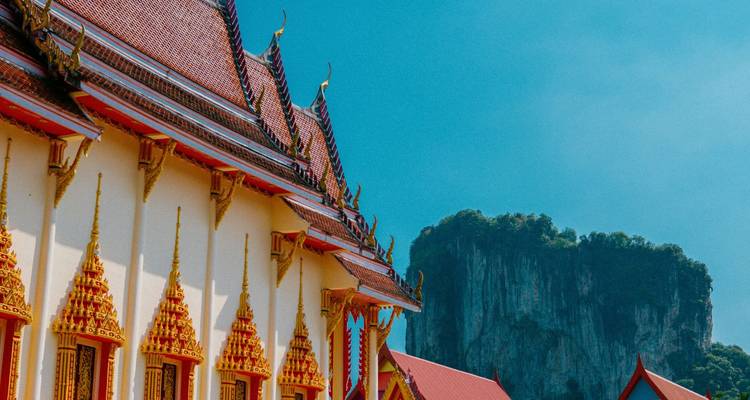 Brightly decorated Thai-style temple roof set against a sheer limestone cliff and blue sky.