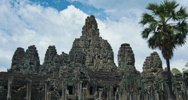 Weathered stone towers of Bayon Temple rising beneath a palm tree and dramatic clouds.