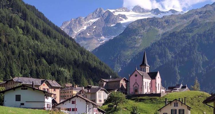 Village pittoresque suisse avec un clocher d'église rose se dressant sur fond de pentes boisées et de pics enneigés.