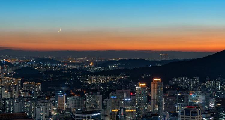 Schemering panorama van Seoul met een wassende maan boven een bergachtige skyline die gloeit in warme tinten.