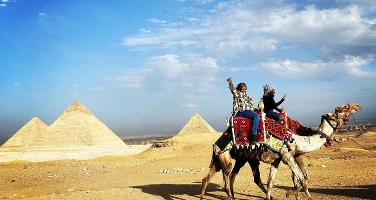 Dos viajeros montan un camello decorado con las tres pirámides de Giza elevándose detrás de ellos bajo un cielo azul.