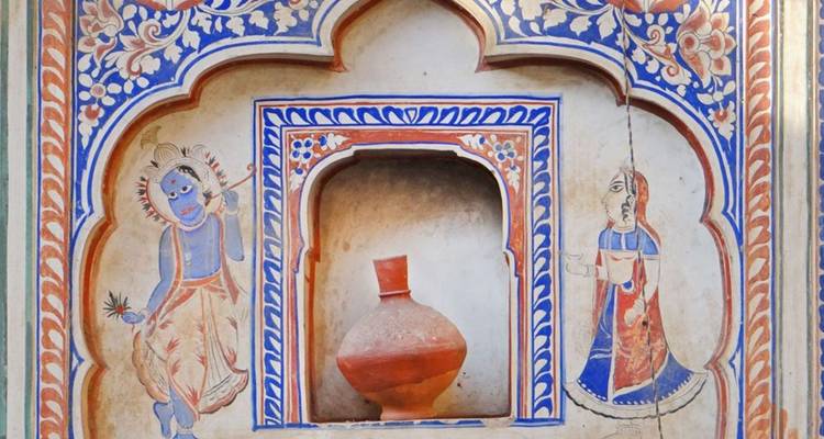 A colorful Rajasthani fresco panel depicts deities and floral patterns surrounding a recessed niche with a clay pot.