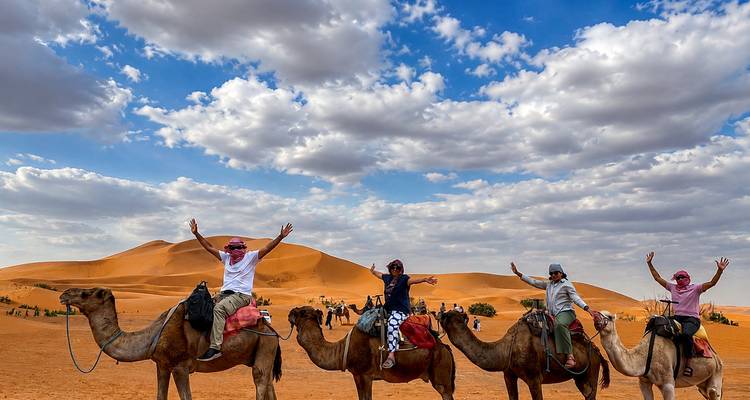 Four riders on camels raise their arms triumphantly against towering orange Erg Chebbi dunes and dramatic clouds.