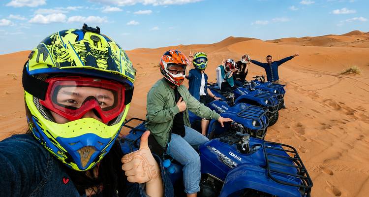 Adventurers in helmets sit on blue ATVs giving thumbs up amid the red sand dunes.
