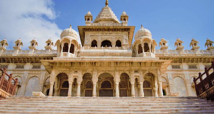 White marble cenotaph with domes and lattice windows rising above broad stone steps under blue sky.