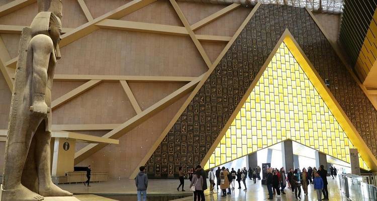 Visitors wander a vast atrium where a colossal pharaonic statue stands before a dramatic triangular wall.