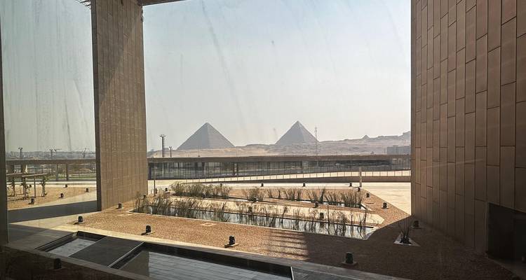 Framed view through vast windows of the museum toward two distant pyramids across the desert.
