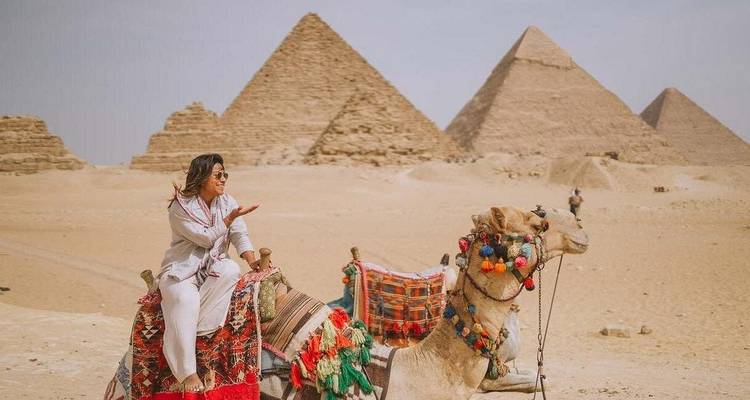 Woman blows a kiss while seated on a decorated camel with the pyramids lined up behind.
