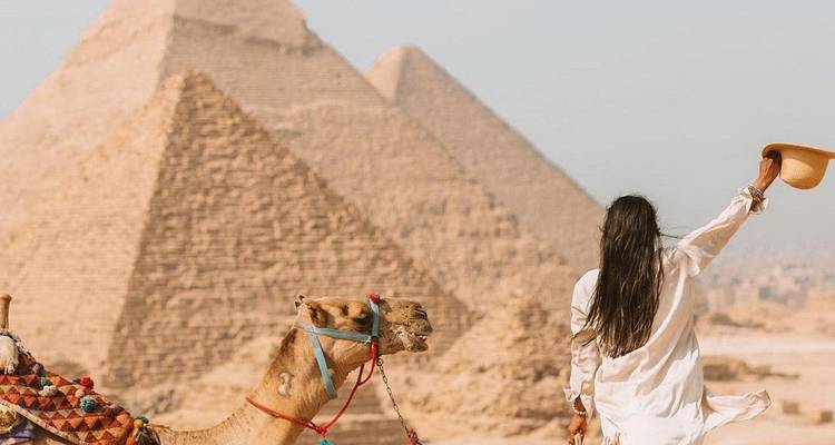 Traveller waves a hat from a camel’s back toward the ancient pyramids.