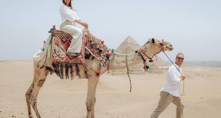 Woman rides high on a camel as her partner leads the reins with pyramids rising behind.
