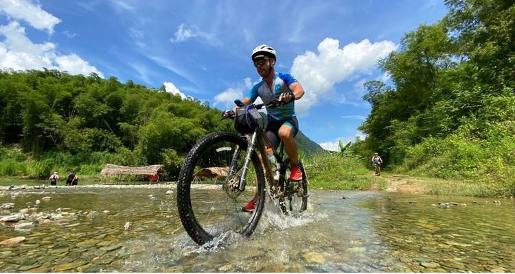 A mountain biker splashes through a shallow creek surrounded by lush forest under a bright blue sky.
