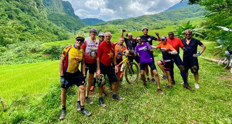 A cheerful group of cyclists pose with their bikes amid vivid green rice terraces and mountain scenery.