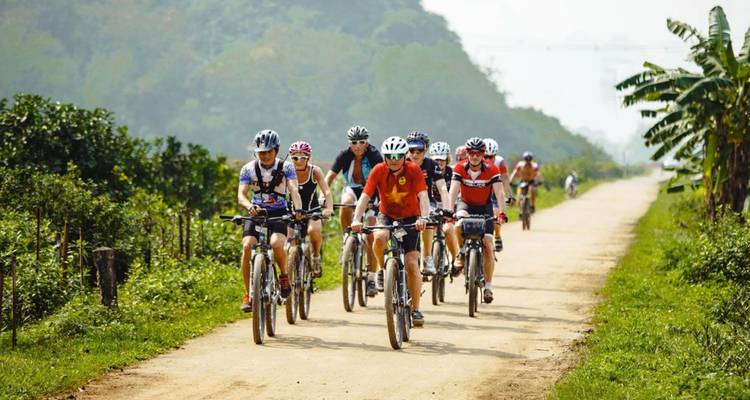 A line of cyclists ride a dusty rural track flanked by tropical vegetation and distant hills.