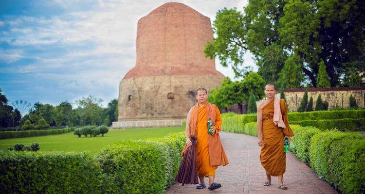 Zwei buddhistische Mönche in safranfarbenen Roben gehen auf einem Ziegelpflasterweg auf die imposante Dhamek Stupa zu, umgeben von gepflegten Gärten.