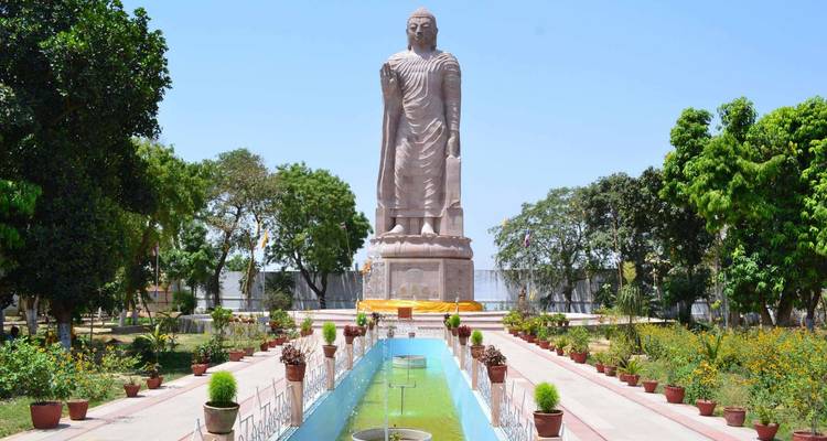 Eine massive stehende Buddha-Statue erhebt sich über einen angelegten Gehweg und einen Springbrunnen an einem hellen Tag.