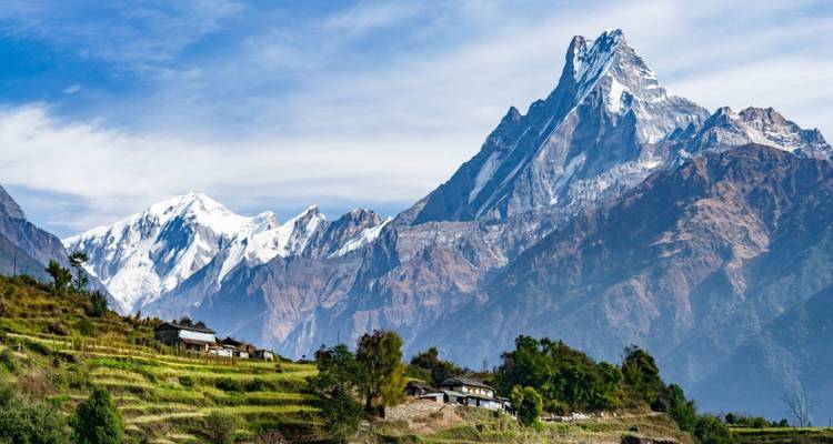 Snow-capped Machhapuchhre towers above terraced fields and small village houses under a bright sky.