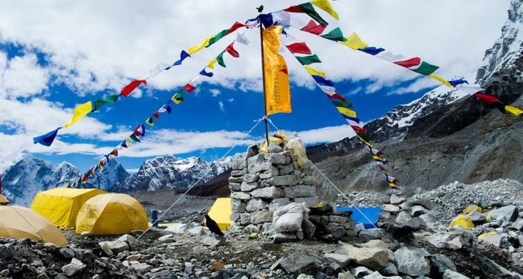 Colorful prayer flags radiate from a stone cairn at a high-altitude Himalayan base camp surrounded by glaciers.