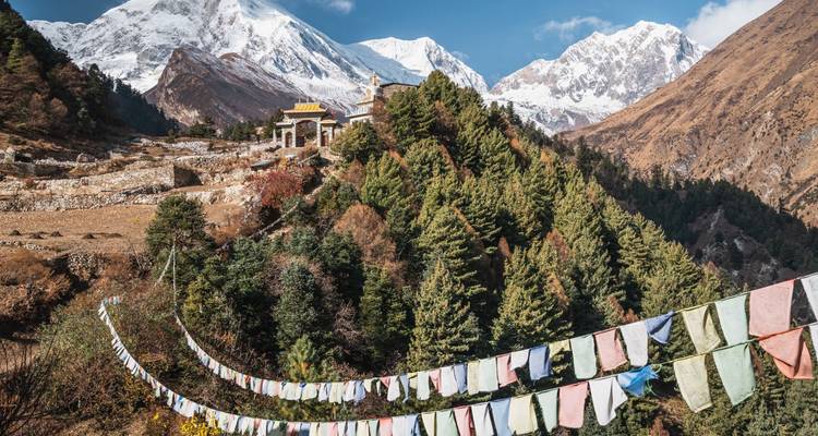 Prayer flags stretch across a verdant Himalayan hillside with snow peaks rising behind a small temple.