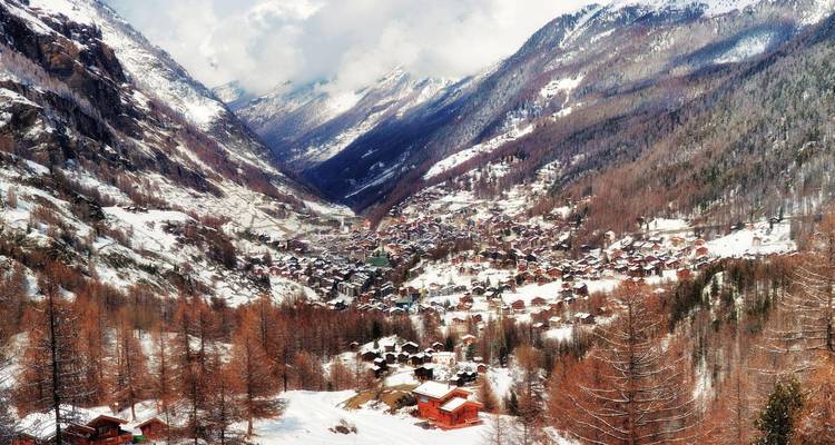 Une vallée alpine enneigée parsemée de chalets s'étend sous des sommets enveloppés de nuages en hiver.