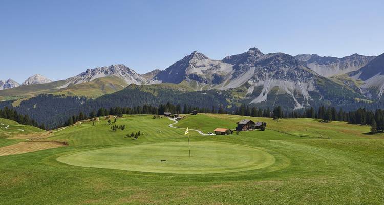 Un green de golf en haute altitude surplombe des pâturages alpins ondulants et des pics gris dentelés sous un ciel dégagé.