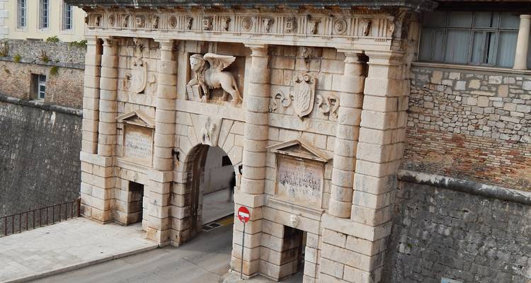 Imposing stone Renaissance gate with carved winged lion and shield motifs guarding a narrow roadway.
