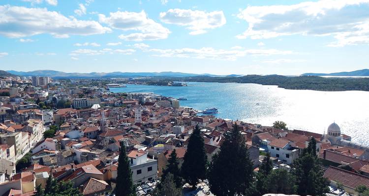 Expansive view over a coastal city, harbor and glittering sea with scattered clouds and mountains beyond.