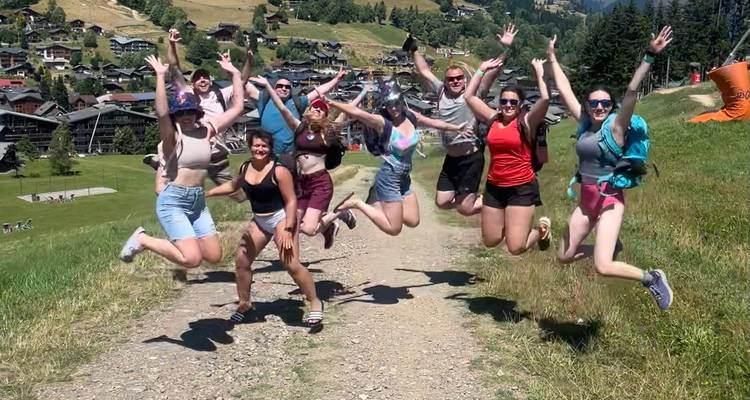 Fröhliche Gruppe, die gleichzeitig auf einem Bergpfad springt, mit einem Dorf darunter.