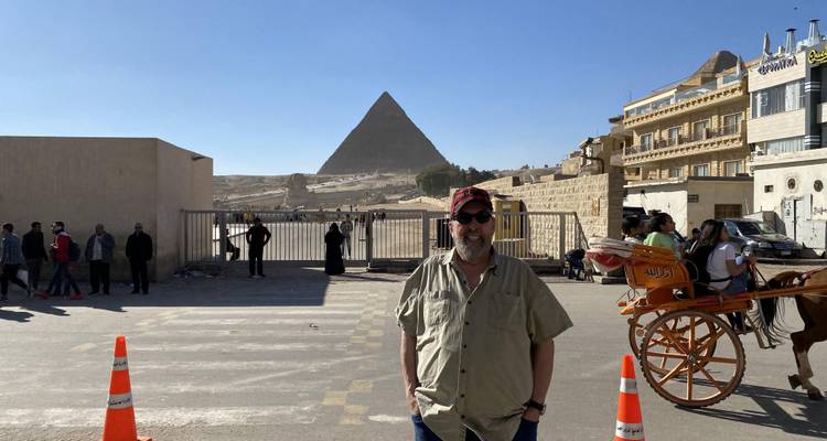 Bezoeker met pet poseert bij de ingang van de straat met pyramidesilhouet in het laat middaglicht.