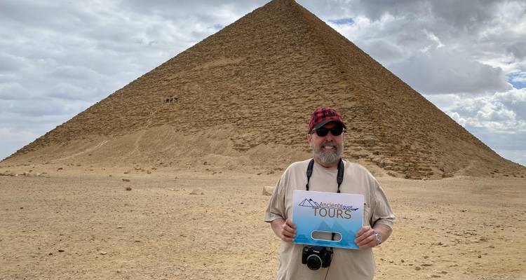 Man met camera staat voor de Knikkende Piramide tegen een dramatische bewolkte lucht.