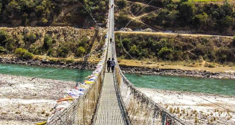 Travellers walk across a long metal suspension bridge high above a turquoise river and rocky banks.