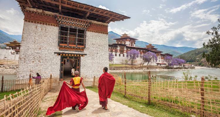 Monks in red robes walk beside a white-stone riverside dzong framed by purple jacaranda trees and mountains.