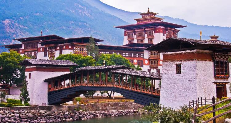 The grand Punakha Dzong and its traditional covered bridge stand against forested hills and a hazy mountain backdrop.