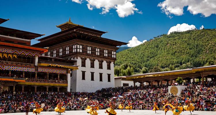 Large crowds watch costumed dancers performing in the courtyard of a Bhutanese monastery against green hills.