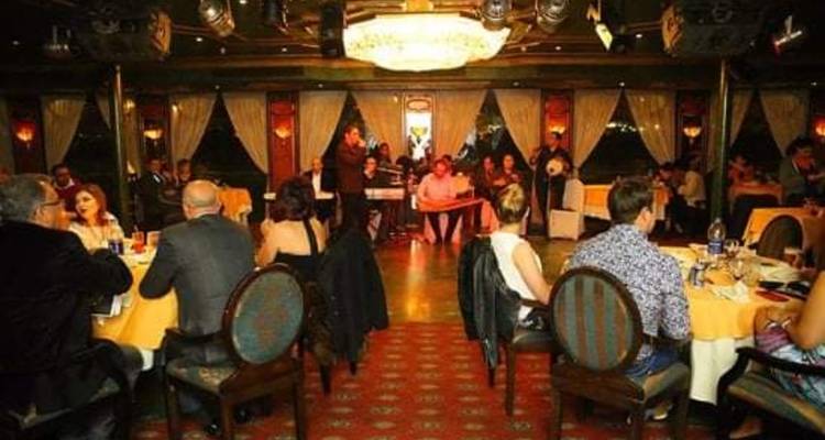 Salon de croisière-dîner avec des convives assis à des tables rondes regardant un spectacle en direct sur scène.