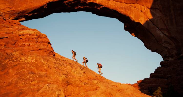 Three hikers ascending beneath a massive natural rock arch at Arches National Park