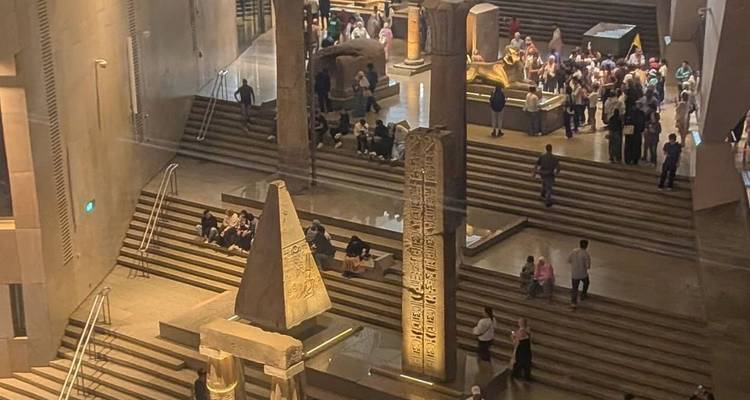 Hall intérieur animé du Grand Musée égyptien avec des foules circulant autour de statues et d'artefacts sur des marches étagées.