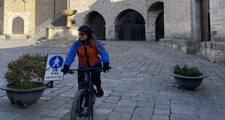 Woman on an e-bike rides through a historic piazza with stone arches and fountain.