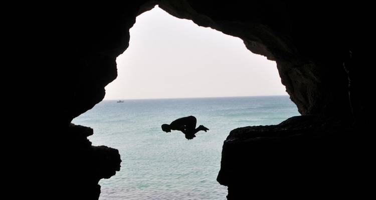 Silhouette of a person mid-flip against the sea as viewed from inside a rocky coastal cave.