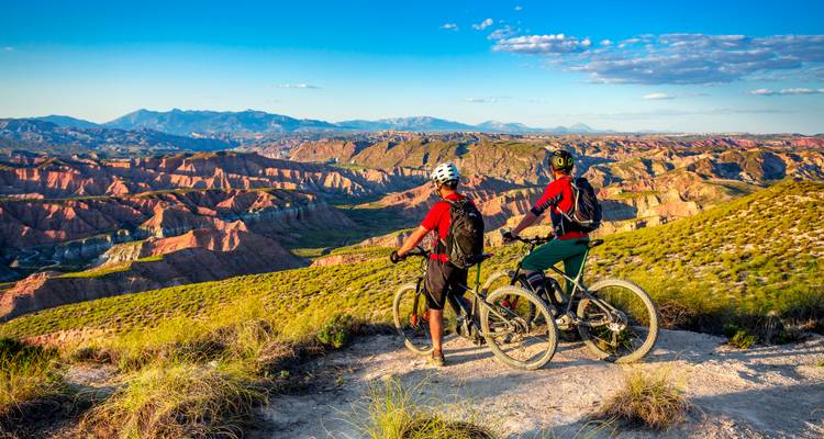 Zwei Mountainbiker machen eine Pause auf einem hohen Bergrücken mit Blick auf farbenfrohe Badlands und entfernte Bergketten